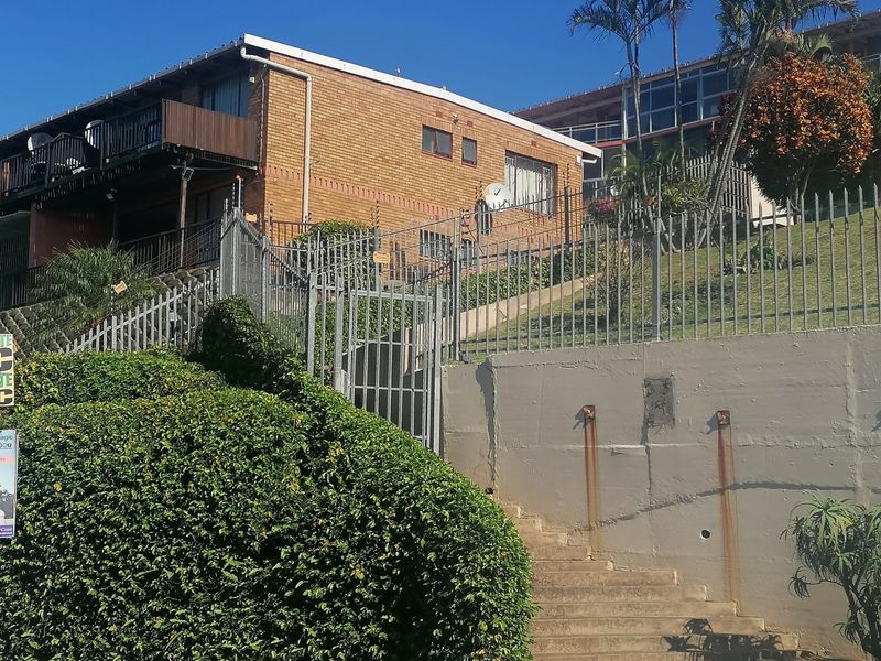 Exterior view of a brick apartment building featuring a concrete retaining wall, security fencing, and a staircase leading to the entrance.