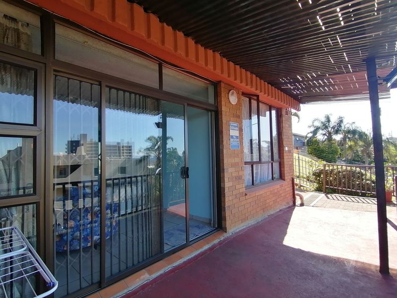 A covered patio area featuring brick exterior walls, sliding glass doors with security gates, and a slatted roof structure.