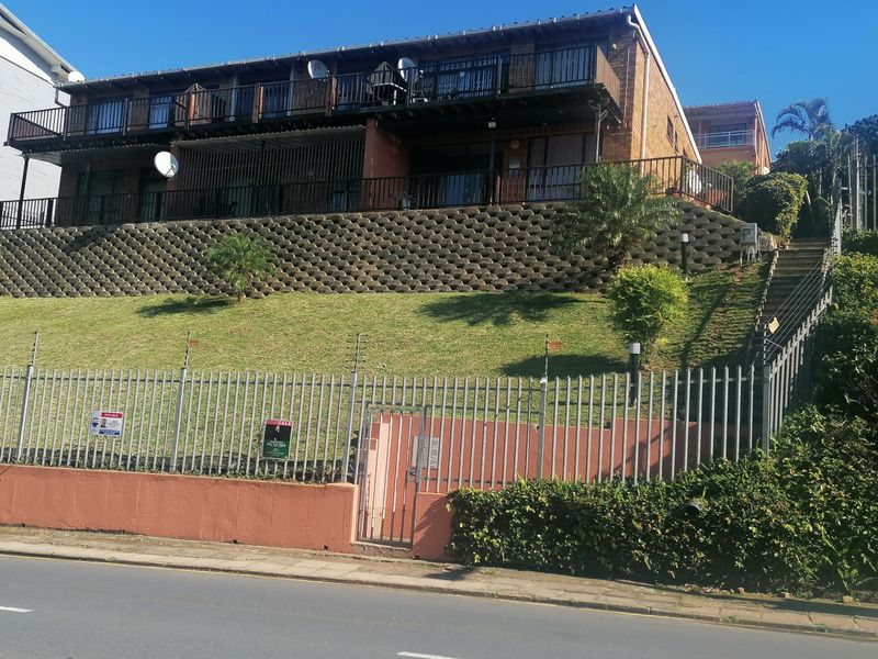 Front exterior view of a multi-story apartment building featuring a prominent retaining wall, security fencing, and balconies.