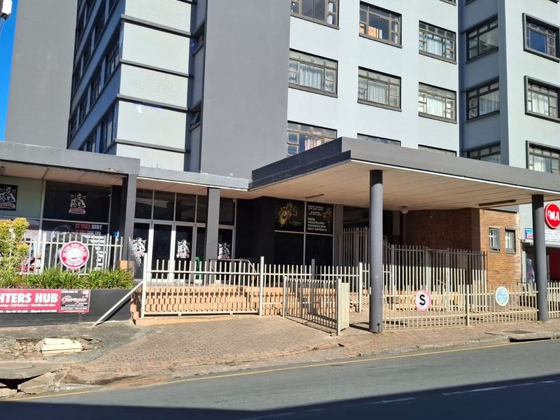 Front exterior view of a multi-story apartment building featuring a covered entrance, paved driveway, and metal security fencing.