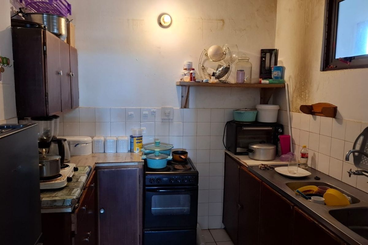 A compact kitchen featuring dark cabinetry, a stainless steel sink, tiled backsplash, and a freestanding stove, requiring cosmetic updates to improve its condition.