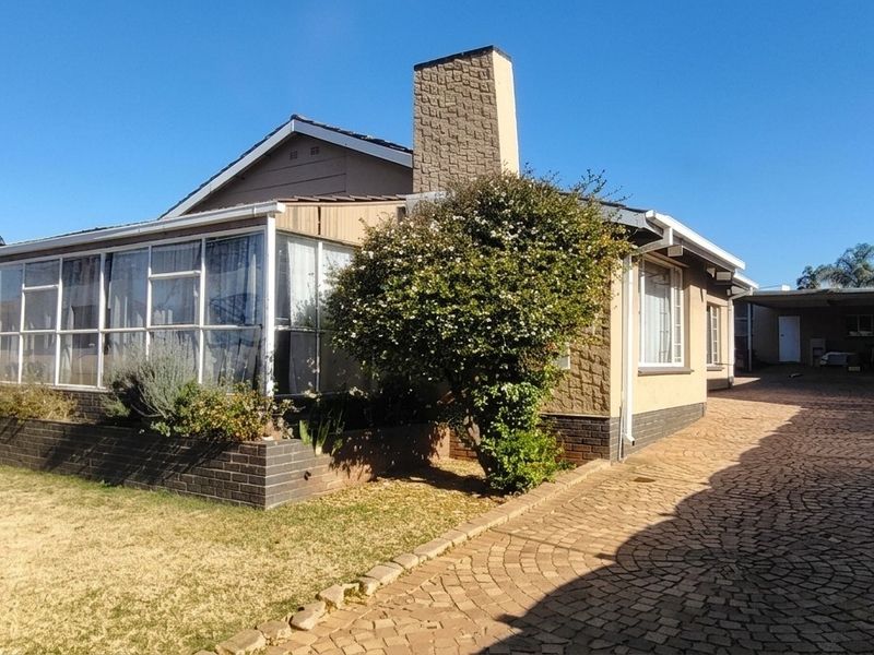 Front view of a residential house featuring a paved driveway, enclosed sunroom, brick chimney, and secure boundary wall with electric fencing.