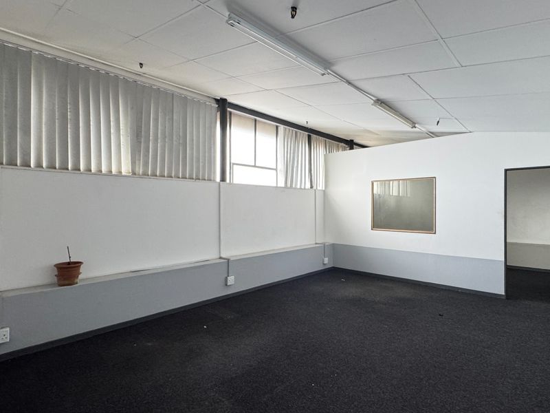 A vacant office space featuring a suspended ceiling, fluorescent lighting, vertical blinds, and a wall-mounted air conditioning unit.