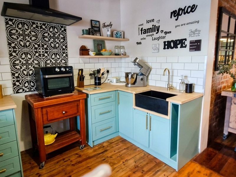 A charming kitchen featuring teal cabinetry, a black apron-front sink, subway tile backsplash, and warm hardwood flooring with an exposed brick accent wall.