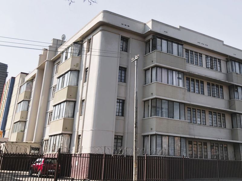 A multi-story apartment building featuring classic architectural lines, steel-framed windows, and secure perimeter fencing with razor wire.