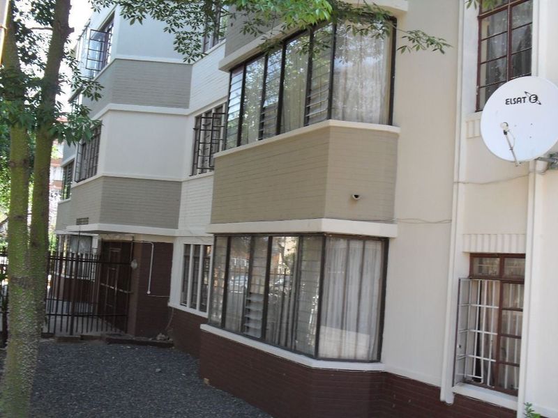 Exterior view of an apartment building featuring multi-level painted brick facade, metal-framed windows with security bars, and a mounted satellite dish.