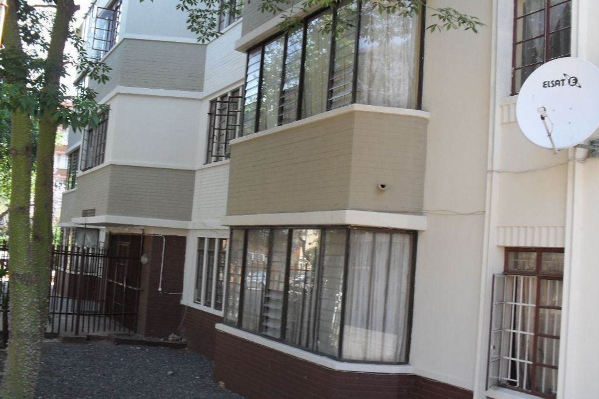 Exterior view of an apartment building featuring multi-level painted brick facade, metal-framed windows with security bars, and a mounted satellite dish.