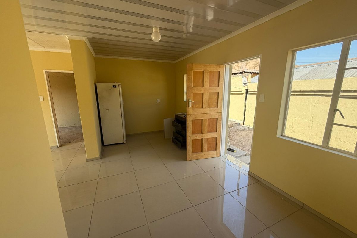 An empty living area featuring tiled flooring, a panelled ceiling, and direct access to the exterior through a timber door.