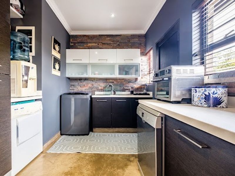 A well-appointed scullery featuring modern dark cabinetry, a stone feature wall, stainless steel sink, and ample storage space with integrated appliance areas.
