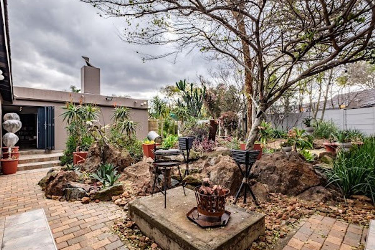 A landscaped rear garden area featuring brick paving, a central concrete plinth, mature trees, and access to the home via folding glass doors.