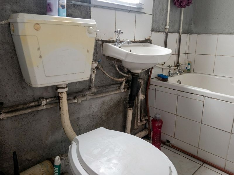 A bathroom featuring a toilet, washbasin, and bathtub with exposed plumbing and dated wall finishes requiring significant renovation and maintenance.