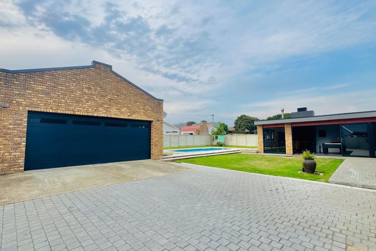Rear exterior view featuring a brick garage, paved driveway, swimming pool, and a covered patio area set within a well-maintained garden.