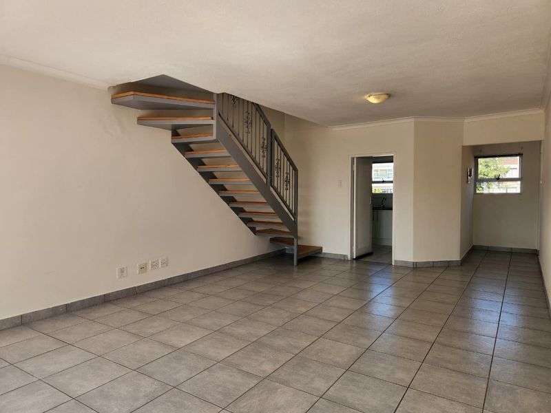 An open-plan living area featuring tiled flooring and a central staircase with a wrought iron balustrade, leading to an adjacent kitchen space.