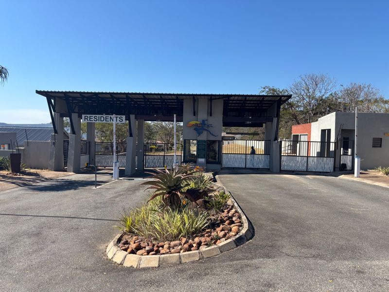 A well-maintained residential estate entrance featuring a secure gated access point, asphalt driveway, and landscaped traffic island.