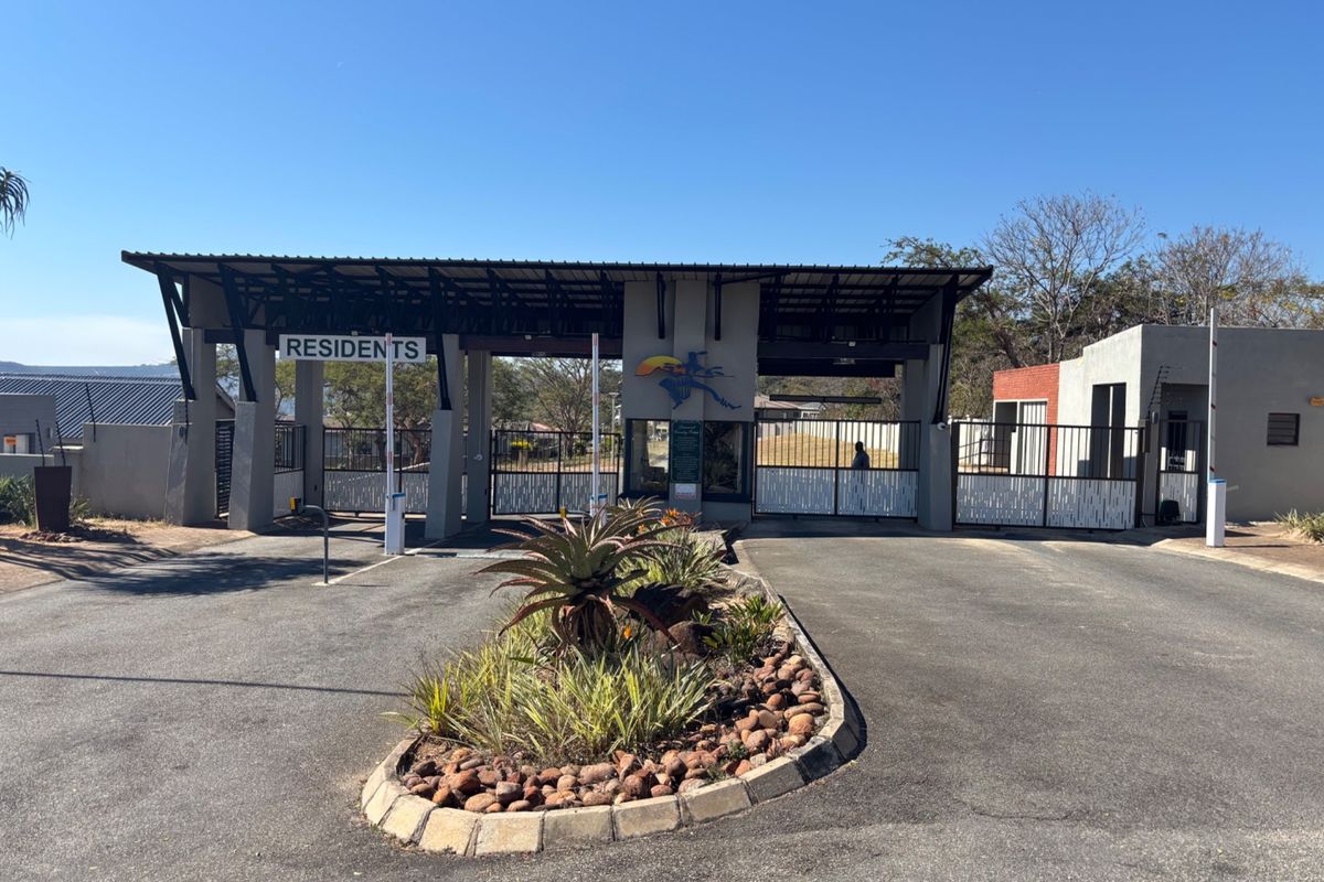 A well-maintained residential estate entrance featuring a secure gated access point, asphalt driveway, and landscaped traffic island.