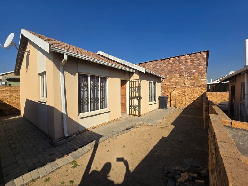 Rear exterior view of a townhouse featuring a paved walkway, security gates, and a private yard area enclosed by brick boundary walls.