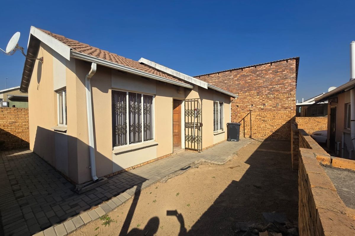 Rear exterior view of a townhouse featuring a paved walkway, security gates, and a private yard area enclosed by brick boundary walls.