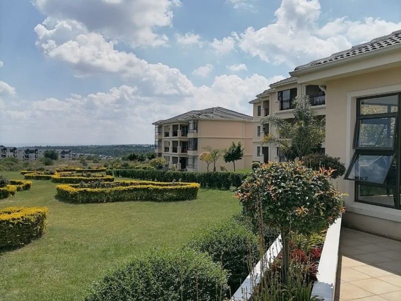 A well-maintained rear exterior view featuring a tiled patio, landscaped garden with manicured hedges, and views of the surrounding apartment complex.