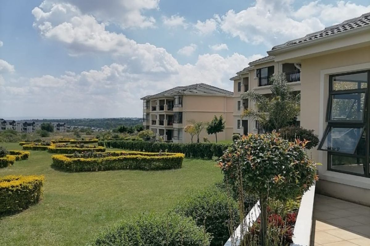 A well-maintained rear exterior view featuring a tiled patio, landscaped garden with manicured hedges, and views of the surrounding apartment complex.