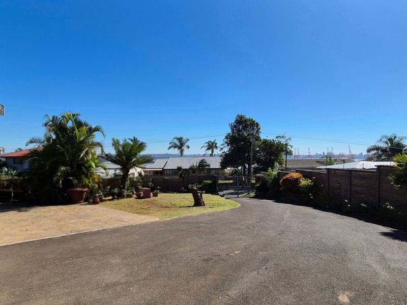 A spacious rear exterior area featuring a paved driveway, established garden, and brick boundary wall with a metal gate.