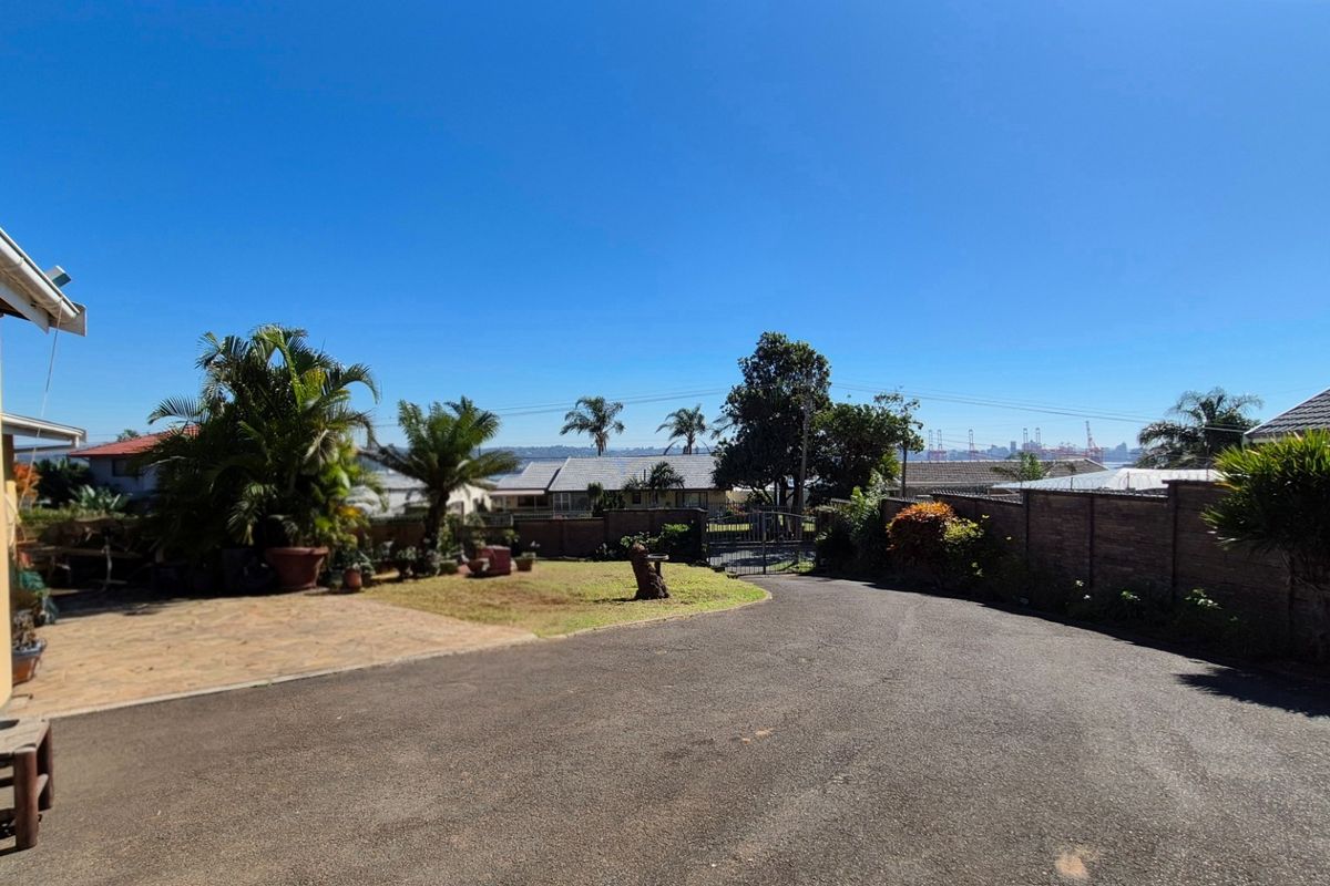 A spacious rear exterior area featuring a paved driveway, established garden, and brick boundary wall with a metal gate.