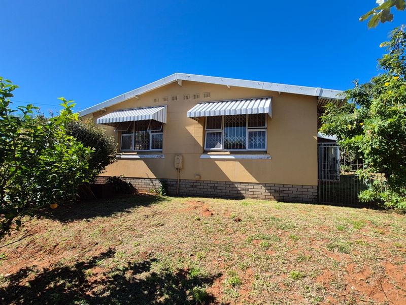 Rear view of the house featuring plastered walls, brick plinth, and metal window awnings with security bars, set against a garden area.