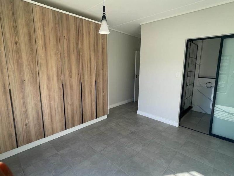 Modern bedroom featuring extensive built-in wood-finish cupboards, neutral tiled flooring, and a sleek sliding door leading to an en-suite bathroom.