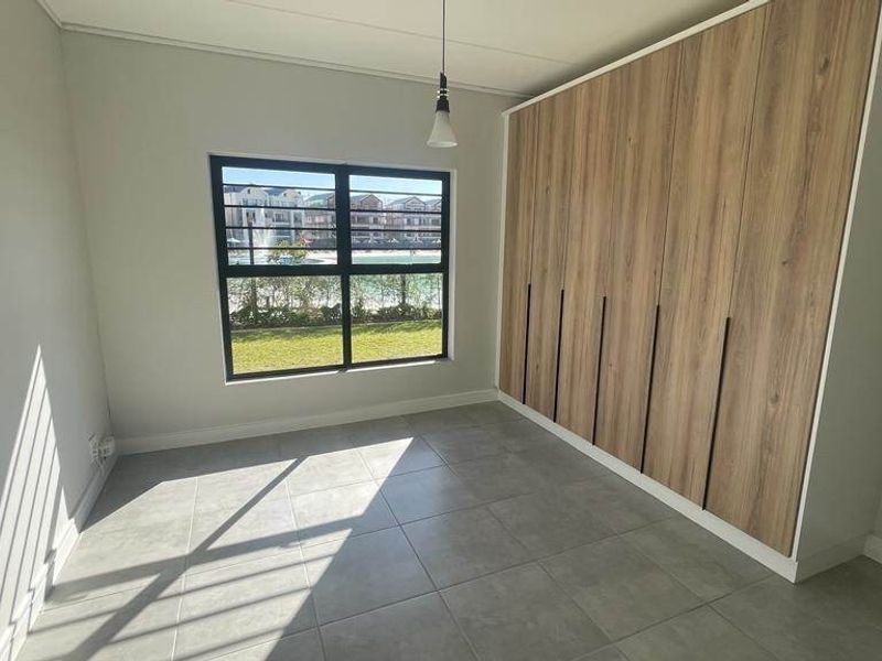 A bright, modern bedroom featuring neutral tiled flooring, extensive built-in wooden cupboards, and a window with security bars overlooking the estate.