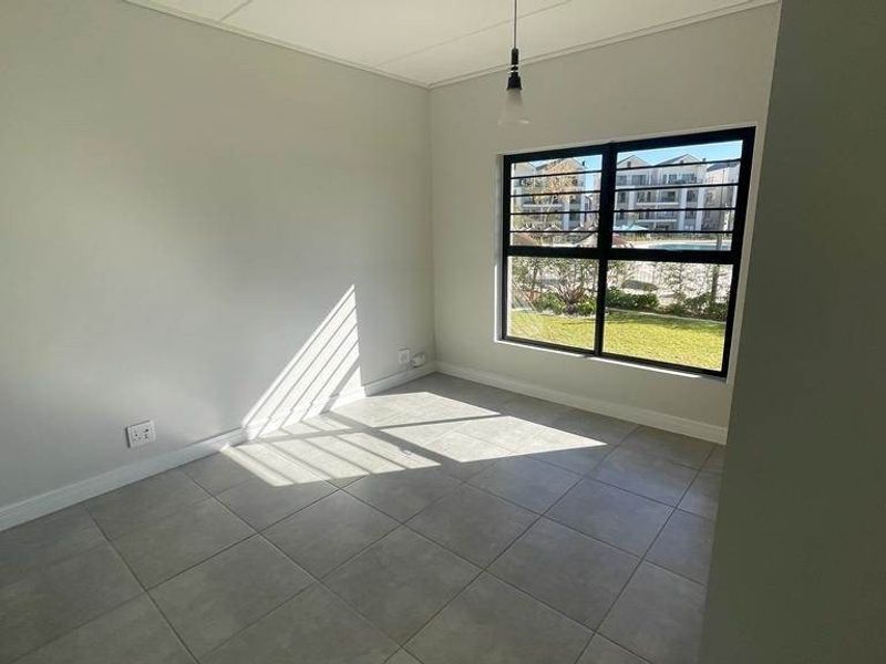 A bright, empty bedroom featuring neutral tiled flooring, clean white walls, and a large window providing natural light and garden views.