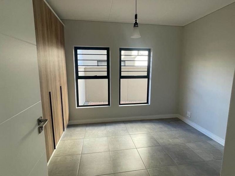 A clean, modern bedroom featuring neutral tiled flooring, built-in wooden cupboards, and large windows with security bars.