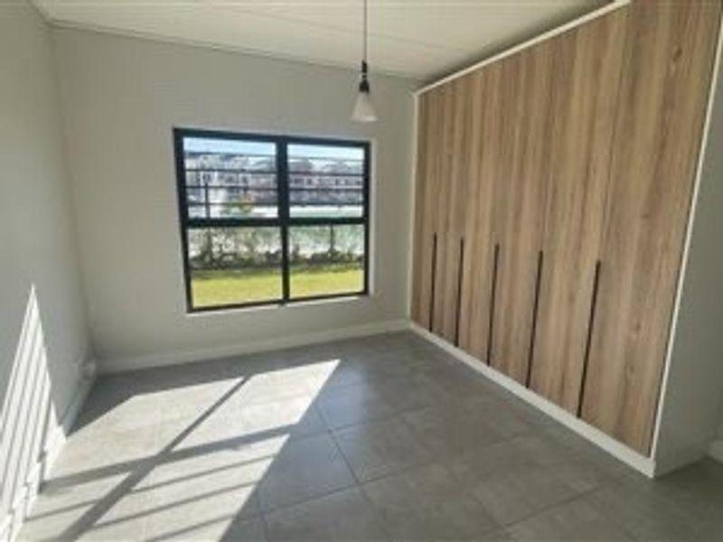 A bright, modern bedroom featuring neutral tiled flooring, a large window, and extensive built-in wooden cabinetry for ample storage.