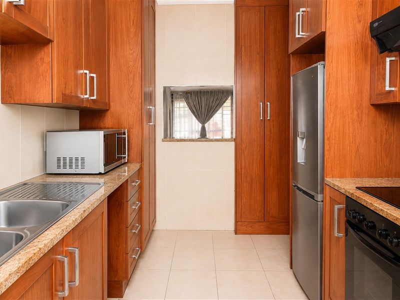 A functional galley-style kitchen featuring warm-toned wooden cabinetry, granite countertops, integrated appliances, and tiled finishes throughout.