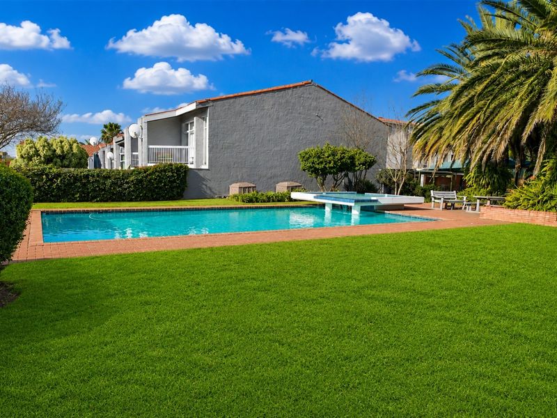 A well-maintained communal swimming pool area featuring a water feature, brick coping, and expansive green lawn within a townhouse complex.
