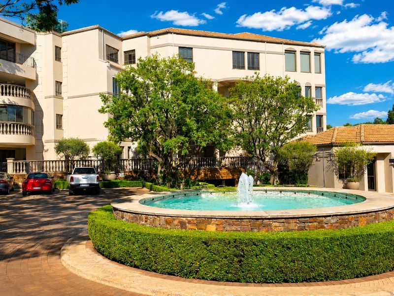 A well-maintained front exterior view featuring a central stone fountain, paved driveway, and elegant multi-story building facade with classic balustrades.