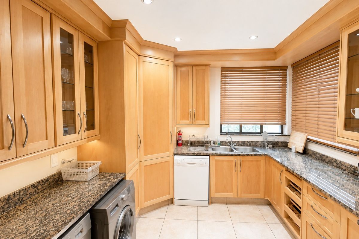 A functional kitchen featuring light wood cabinetry, granite countertops, a double basin sink, and integrated appliances with tiled flooring and natural light.
