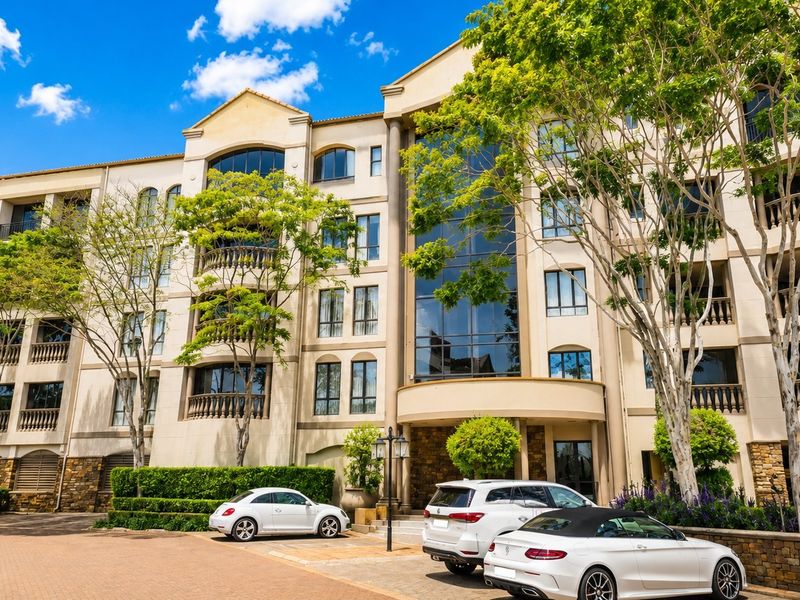 A multi-story residential building facade featuring stone cladding, decorative balustrades, and large glass windows with a paved driveway entrance.