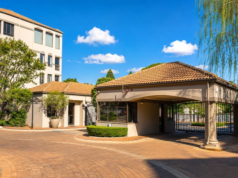A well-maintained security entrance and gatehouse featuring a paved driveway and multi-story residential building in the background.