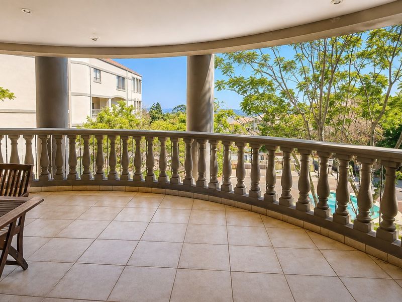 Spacious covered balcony featuring tiled flooring, decorative balustrades, and structural columns, offering an elevated view of the surrounding estate and pool area.