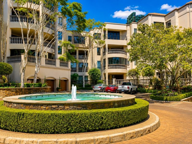 A well-maintained residential complex entrance featuring a central stone fountain, paved driveway, manicured hedges, and elegant architectural balustrades.