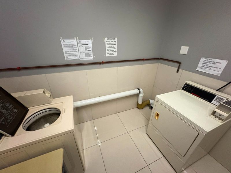 A functional laundry area featuring tiled flooring, tiled splashbacks, and exposed utility pipework for washing machines and dryers.