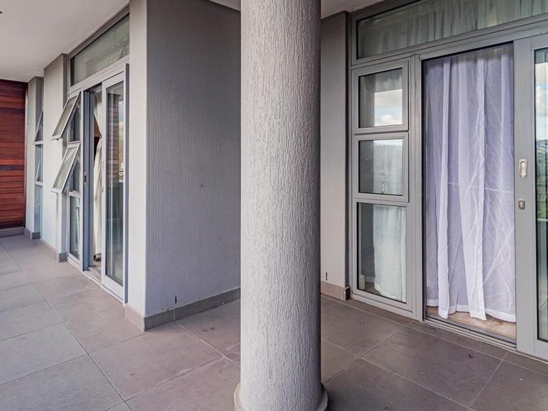 A spacious covered balcony featuring tiled flooring, a central concrete column, and sliding glass door access to the interior living areas.