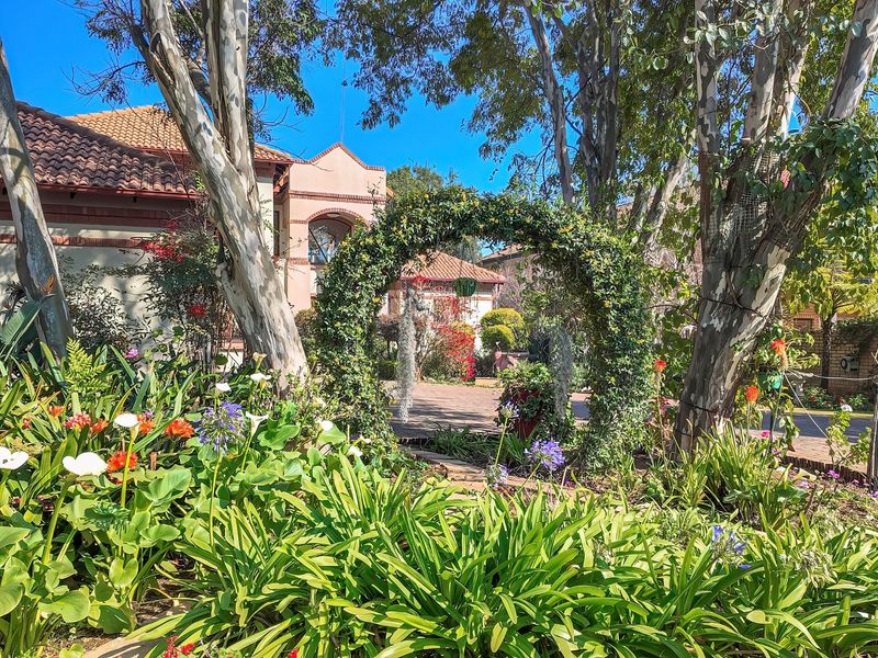 A lush, well-maintained garden featuring mature trees, vibrant flower beds, and a decorative archway leading toward the main house entrance.