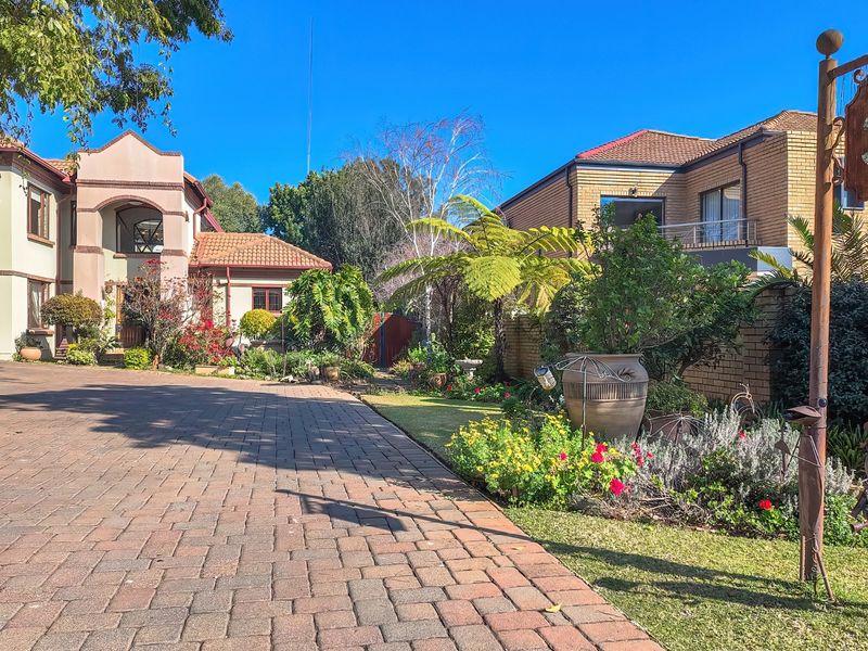 A well-maintained front exterior view featuring a paved driveway, established garden landscaping, and a multi-level brick and rendered residential facade.