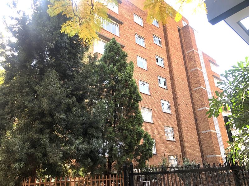 A view of the multi-story brick apartment building facade, featuring established trees and secure perimeter fencing.