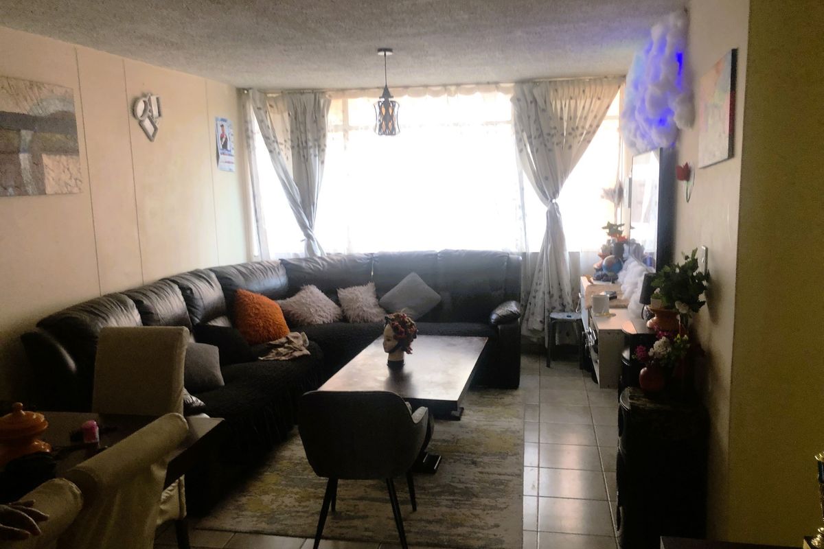 A living room area featuring tiled flooring and a textured ceiling, with natural light entering through a large window.