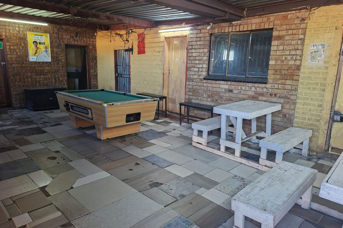 A covered outdoor patio area featuring exposed brick walls, tiled flooring, and a corrugated metal roof, suitable for a casual entertainment or social space.