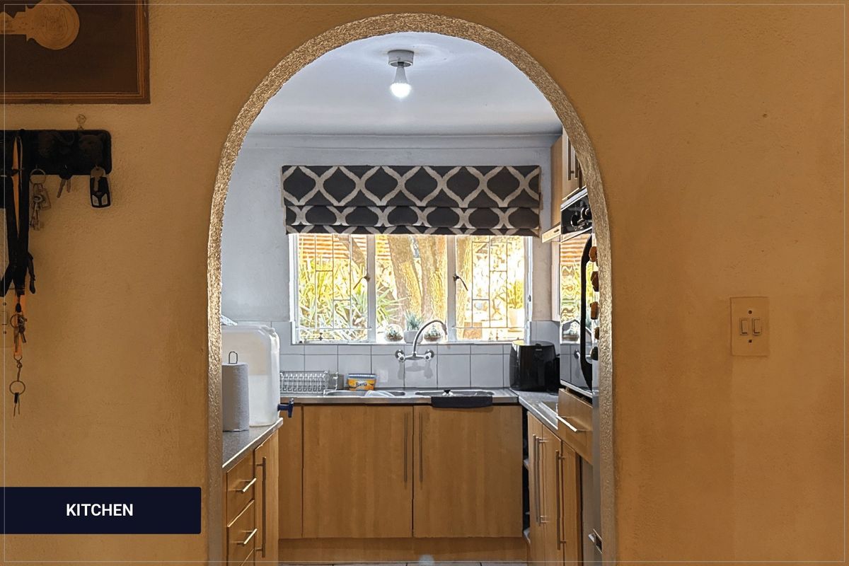 A functional kitchen viewed through an arched doorway, featuring wooden cabinetry, a stainless steel sink, and a tiled backsplash with natural light.