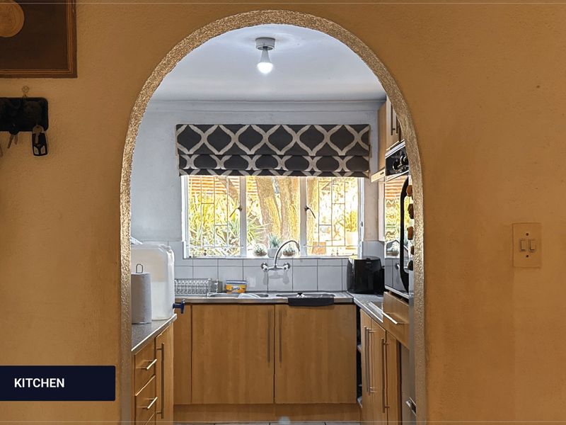 A functional kitchen viewed through an arched doorway, featuring wooden cabinetry, a stainless steel sink, and a tiled backsplash with natural light.