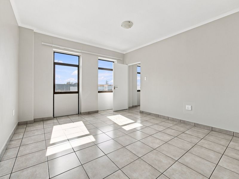 A bright, empty bedroom featuring neutral wall paint, tiled flooring, and large windows providing natural light.