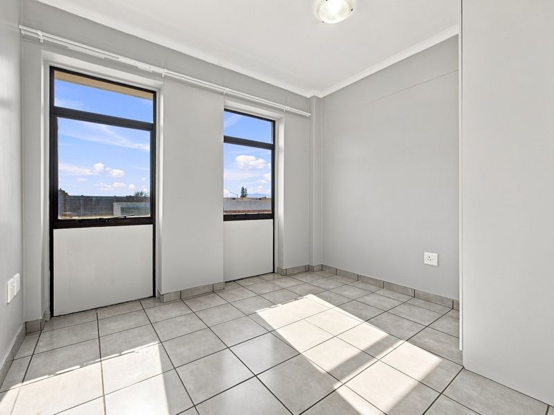 A bright, empty bedroom featuring neutral grey walls, tiled flooring, and large aluminium-framed windows allowing for ample natural light.